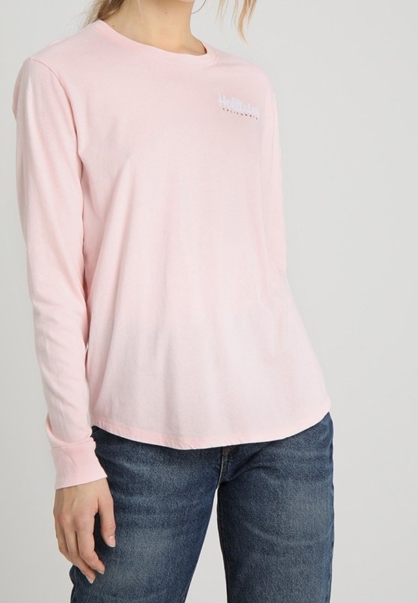 Maglietta a maniche lunghe rosa in morbido cotone, con scollo rotondo e un logo bianco sul petto a sinistra. Abbinata a jeans blu.