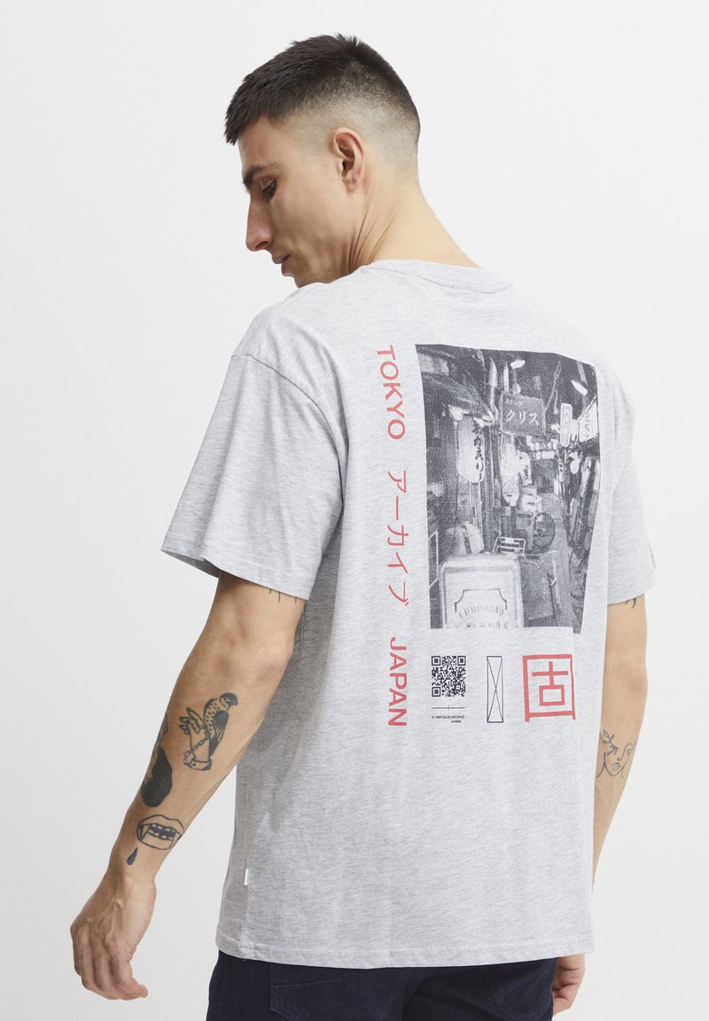 Šedé tričko s krátkým rukávem, na zádech s fotografickým potiskem, červeným japonským textem a QR kódem. Materiál vypadá jako bavlna.