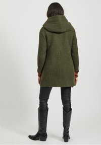 Cappotto verde texturizzato con cappuccio e vestibilità ampia. Maniche lunghe e pantaloni in pelle nera. Stivali neri fino al ginocchio completano l'outfit.