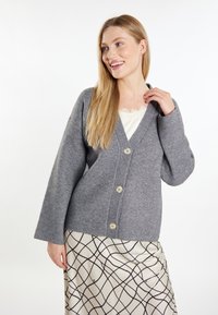 Cardigan tricotat gri cu decolteu în V, mâneci lungi și cinci nasturi mari. Puratat peste o fustă crem cu un model geometric negru.