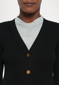 Black ribbed cardigan featuring a deep V-neck, two golden buttons with a logo, layered over a light blue high-neck top.