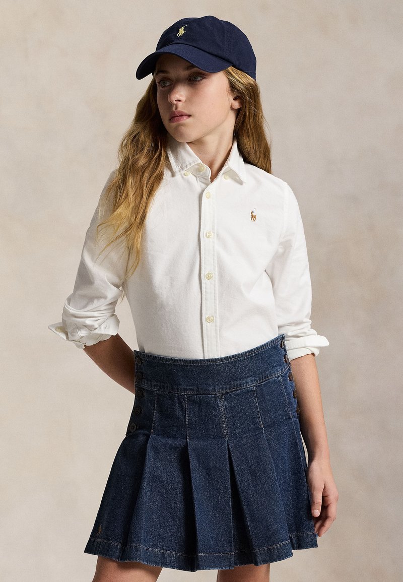 Persona joven que lleva una camisa blanca abotonada, falda vaquera oscura plisada y gorra azul marino con un logo de caballo, mirando hacia un lado.