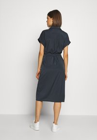 Abito camicia blu navy con maniche corte arrotolate, vita con cintura e orlo fino al ginocchio. Tessuto morbido e due tasche posteriori visibili.