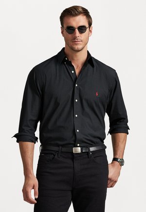 Camisa preta de botão com mangas arregaçadas, apresentando um pequeno logotipo vermelho no peito. Usada com jeans pretos e um cinto prateado. Óculos de sol escuros.