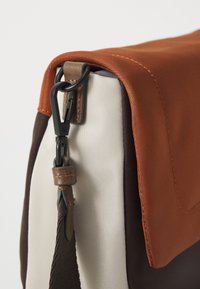 Borsa a tracolla con patta marrone, corpo beige e marrone scuro, dotata di accessori in metallo nero e dettagli in pelle. Materiali texture presenti.