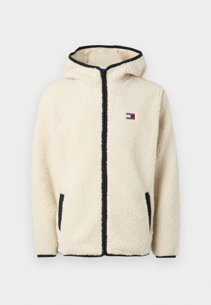Un hoodie en polaire couleur crème avec fermeture éclair sur le devant, bord noir et patch logo. Comprend des poches latérales et un tissu doux et texturé.