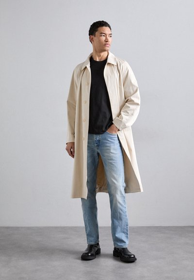 Beige lang frakk med krage, knapper foran, kombinert med en svart skjorte og lyseblå jeans. svarte ankelstøvletter, glatt tekstur.
