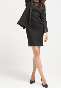 Svart åtsittande klänning med spetskrage och blazer med trekvartsärm, slät textur, knälång, ihop med svarta högklackade skor.