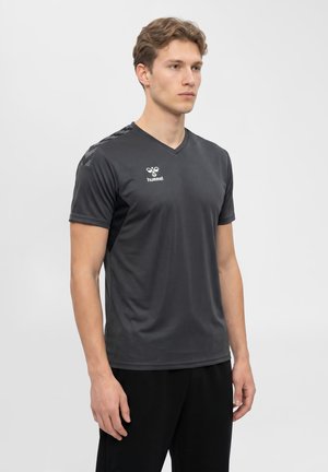 Młody mężczyzna w czarnej sportowej koszulce z krótkim rękawem z logo hummel, stojący na jednolitym jasnoszarym tle.