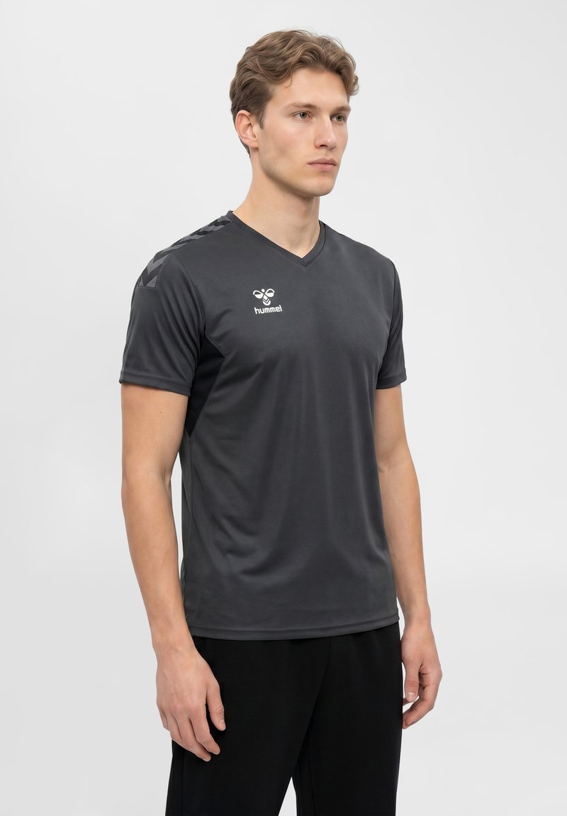 Młody mężczyzna w czarnej sportowej koszulce z krótkim rękawem z logo hummel, stojący na jednolitym jasnoszarym tle.