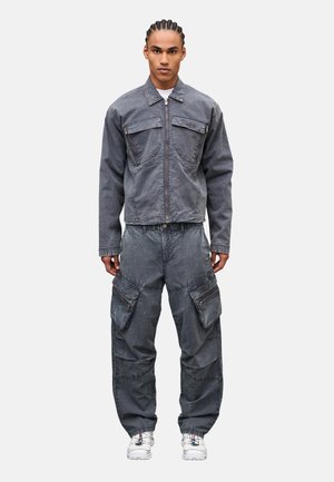 Joven con el cabello trenzado, vestido con una chaqueta gris con cremallera y pantalones cargo a juego, y zapatillas blancas, de pie erguido contra un fondo blanco.