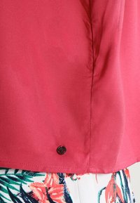 Rotes Stoffoberteil mit einem kleinen runden schwarzen Logo nahe dem Saum, kombiniert mit weißem Stoff, der grüne und orangefarbene Blumenmuster aufweist.