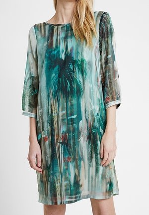 Femme portant une robe mi-longue avec un imprimé abstrait de palmiers et de fleurs en vert et bleu, avec des manches trois-quarts et un col rond.