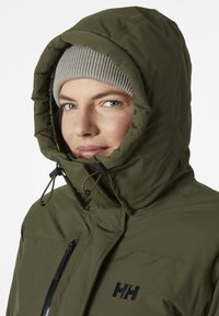 Giacca isolante verde oliva con colletto alto e cappuccio regolabile, dotata di una tasca con zip e logo nero sul petto.