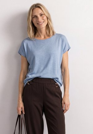 Femme portant un haut bleu clair à manches courtes et un pantalon taille haute foncé, souriante et tenant un sac à main marron foncé à ses côtés.