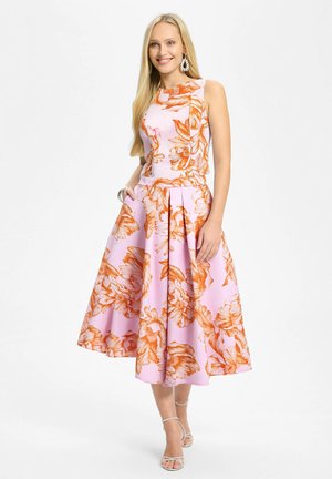 Smilende kvinde iført ærmeløs lyserød midi-kjole med stort orange blomsterprint, sølvfarvede creoler, armbånd og hvide sandaletter med hæl.