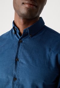 Gros plan sur une personne portant une chemise en velours côtelé bleu boutonnée avec un col boutonné, sur un fond uni.