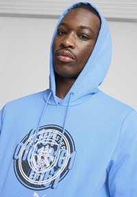 Svijetloplavi dukserica s grafikom sprijeda koja prikazuje "Manchester City" u crnoj boji, s vezicama i teksturiranim materijalom.