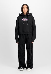 Felpa nera corta con graphic text bianco e rosa, tasca frontale, lacci bianchi, abbinata a pantaloni neri larghi e sneakers bianche.