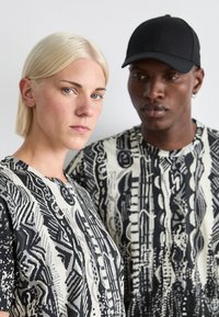 Twee modellen dragen losvallende, gemusterde tops in zwart en wit, met abstracte ontwerpen en texturen. Eén model draagt een zwarte pet.