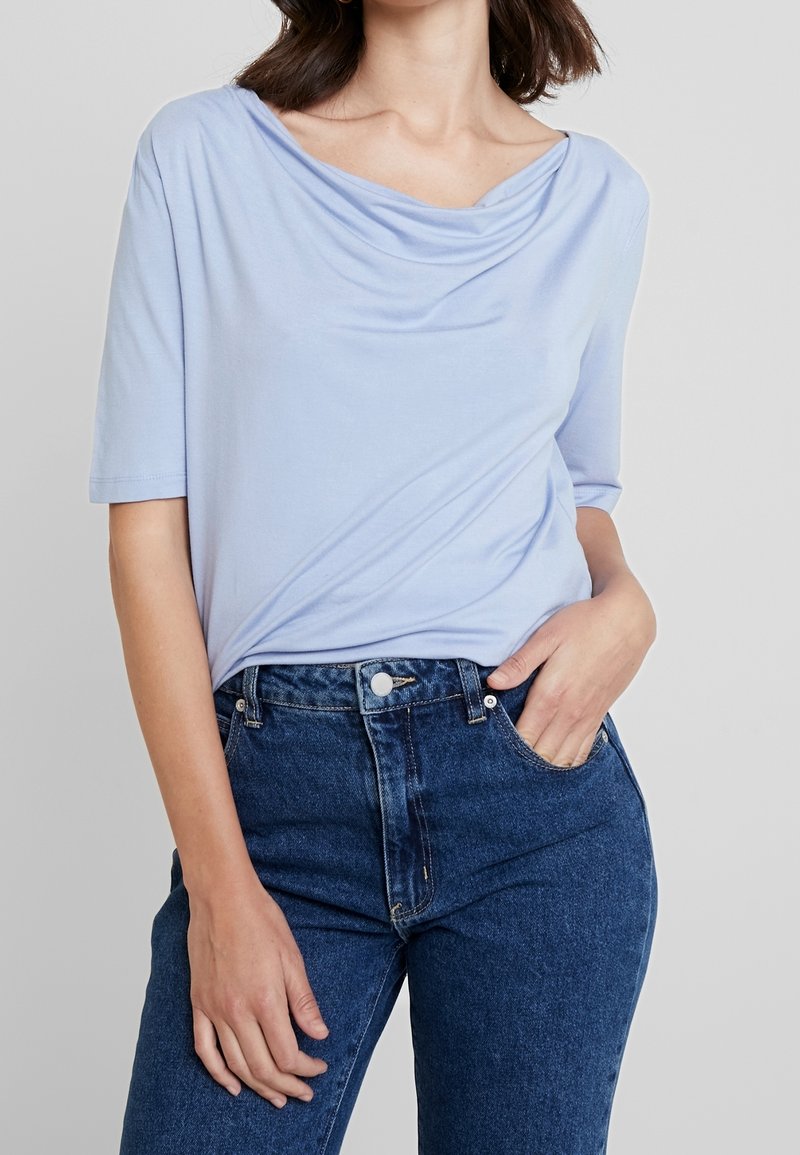 Femme portant un haut à manches courtes bleu clair à col drapé et un jean taille haute bleu foncé, une main dans la poche.
