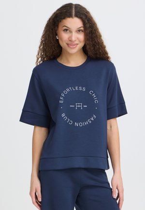 Žena s kudrnatými vlasy, která má na sobě tmavě modré tričko a kalhoty, tričko má text „EFFORTLESS CHIC FASHION CLUB“ v kruhovém designu.