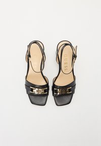 Zwarte leren slingback hakken met vierkante neus, voorzien van gouden hardwareaccenten en een ontwerp met een bandje. Lichte tan binnenzool toont de merknaam "GUESS."