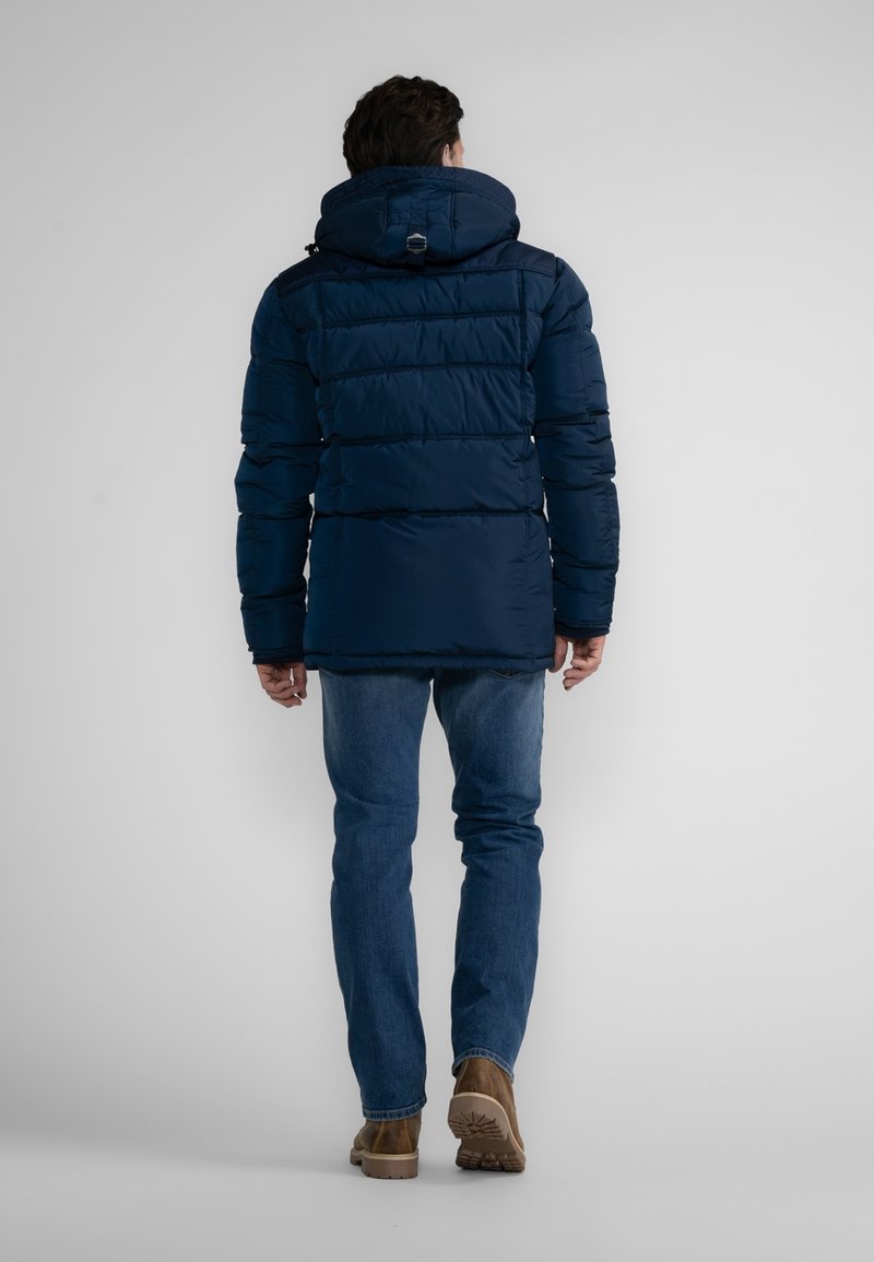 Veste rembourrée bleu marine avec une capuche, dotée de coutures horizontales, de poches zippées et de poignets réglables. Portée avec un jean bleu et des bottes marron.