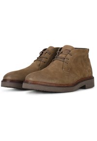 Botines de ante marrón con punta redonda, minimalista en el sistema de cordones y una suela exterior marrón en contraste. Presenta una superficie texturizada y acentos cosidos.