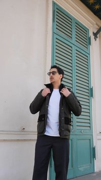 Homme ajustant le col d'une veste sombre sur une chemise blanche, debout devant de grandes portes à volets bleu-vert sur un mur beige.