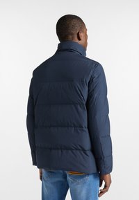 Giacca trapuntata blu navy con colletto alto, design a quadri e superficie liscia. Indossata sopra jeans azzurri.
