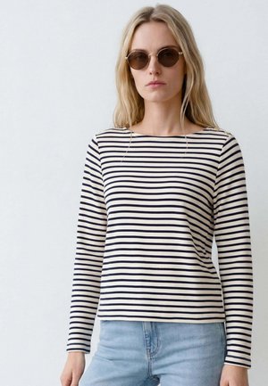 Frau mit langen blonden Haaren, die runde Sonnenbrillen, ein schwarz-weiß gestreiftes Langarmshirt und hellblaue Jeans vor einem einfarbigen Hintergrund trägt.