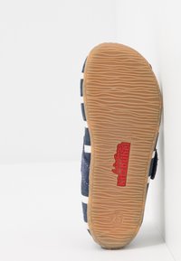 Kengät, joissa on raidoitettu sinivalkoinen kanvaspinta ja kumipohja, jossa on teksturoitu vaaleanruskea pinta, jolla on punainen logo. Koko 25.