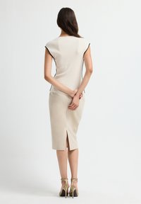 Vrouw staat met de handen gekruist achter de rug, gekleed in een crèmekleurige mouwloze top met zwarte randen, een beige midi rok met een split aan de achterkant en beige hoge hakken.