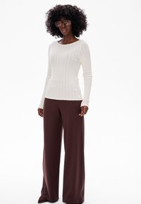 Biała, prążkowana bluzka z długim rękawem i okrągłym dekoltem; zestawiona z szerokimi brązowymi spodniami z rozcięciami po bokach przy kostkach. Duże okulary.