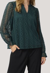 Blouse bleu sarcelle à motifs avec manches en dentelle et encolure en V. Dispose d'une surface texturée avec des motifs géométriques et des manches longues légèrement bouffantes.