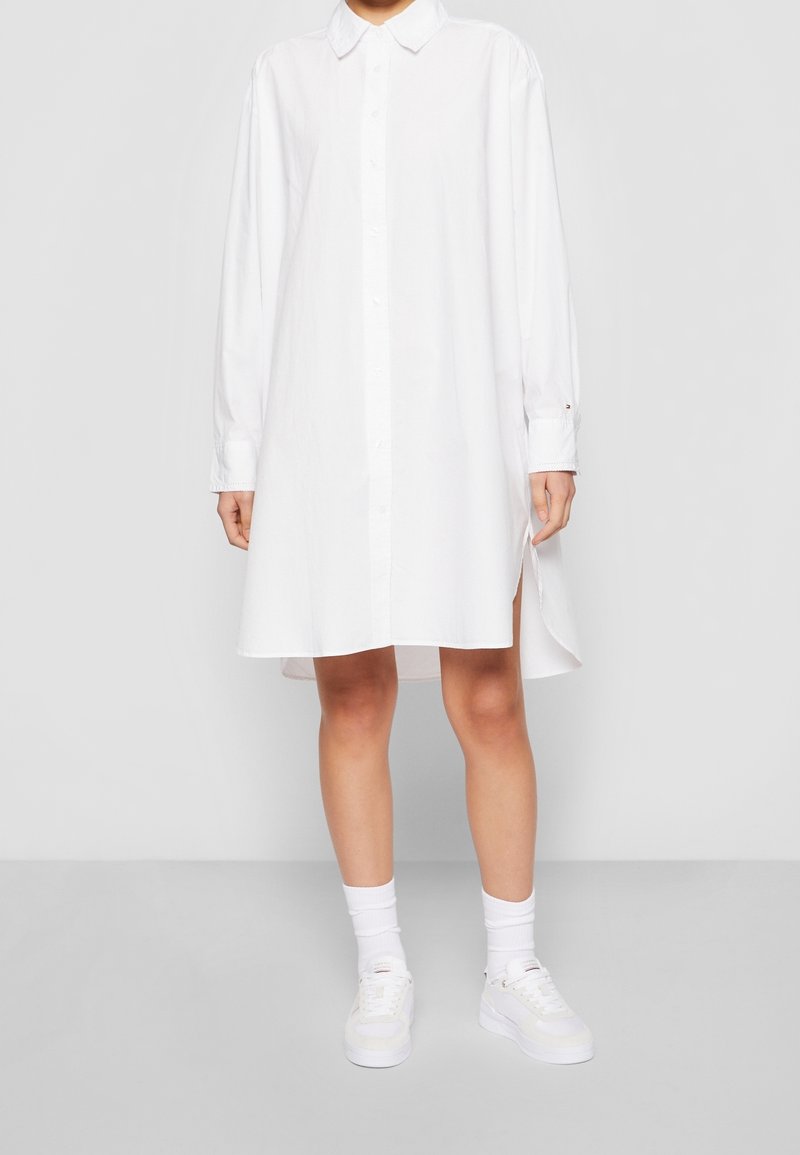 Chemise blanche à manches longues, avec fentes sur les côtés et col classique ; fabriquée en tissu léger avec une texture lisse.