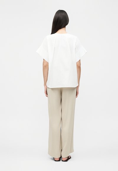 Γυναίκα με μακριά σκούρα μαλλιά στέκεται με την πλάτη γυρισμένη, φοράει άσπρο, άνετο τοπ με κοντά μανίκια, μπεζ παντελόνια φαρδιάς γραμμής και μαύρα σανδάλια σε απλό φόντο.