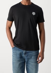 Black cotton t-shirt featuring a small embroidered tiger logo on the chest, round neckline, and short sleeves, paired with light blue jeans.