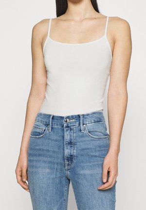 Vrouw draagt een aansluitend wit spaghettihemdje, gestopt in een high-waisted blauwe spijkerbroek met knoop- en ritssluiting.