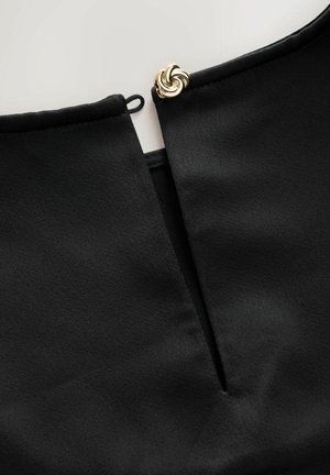 Detailní záběr na černý výstřih oděvu s otvorem ve tvaru klíče a ozdobným zlatým knoflíkem ve tvaru splétané uzlové vazby.