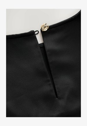 Lähikuva mustasta vaatteesta, jossa on avaus kulkukanavalle ja koristeellinen kultainen kierre solki.