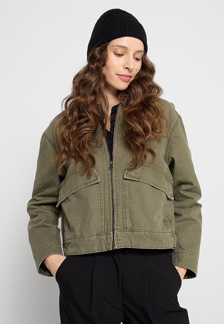 Oliwkowa krótka kurtka z zamek błyskawicznym, dwiema przednimi kieszeniami i mocnym materiałem. Zestawiona z czarną dzianinową czapką i czarną bluzką.