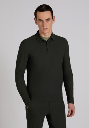 Młody mężczyzna o ciemnych włosach, ubrany w czarną koszulkę polo z długim rękawem i pasujące spodnie, stojący z jedną ręką w kieszeni.