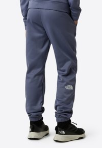 Plave sportske hlače s elastičnim manžetama i malim bijelim logom na bedro, uparene s crnim sportskim tenisicama koje imaju bijeli detalj.