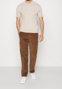 Beige kortärmad stickad polo skjorta med ett strukturerat mönster, tillsammans med bruna kordbyxor och vita sneakers, stående framför en enkel bakgrund.