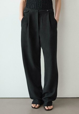 Persona che indossa pantaloni neri larghi con pieghe e bottoni sul davanti, mani in tasca, abbinati a sandali neri aperti sulla punta su pavimento grigio.