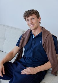 Marinblå polo skjorta, brun tröja som hänger över axlarna, mörka jeans, sitter på en ljusbeige soffa, naturligt ljus, neutral bakgrund.