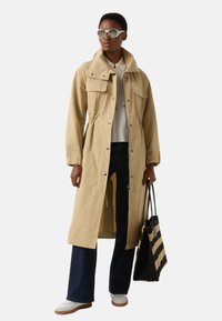 Beżowy długi płaszcz z dwoma przednimi kieszeniami, sznurkiem w talii i zatrzaskami. Połączony z ciemnoniebieskimi, rozszerzanymi dżinsami i białymi tenisówkami.