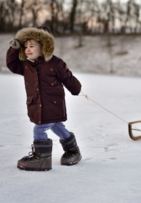 Vaikas dėvintis rudą parką su kailiniu gaubtu, mūvintis mėlynas džinsines kelnes ir juodus žieminius batus, traukia medinę rogutę per sniegą.